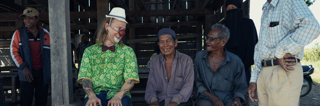 En clown i röd näsa, hatt, fluga och grönmönstrad skjorta sitter bredvid två äldre herrar. De tittar på varandra och skrattar.