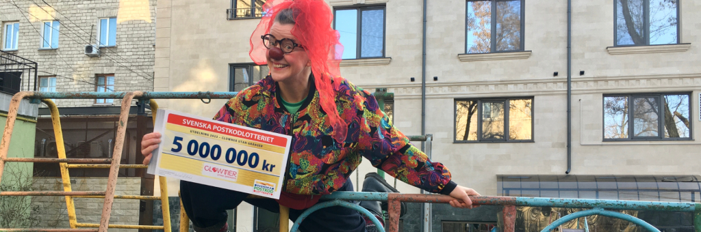 En clown med röd näsa ler mot kameran och håller i en check med summan 5 miljoner kronor