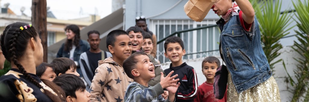 Flera barn står runt en artist och tittar på ett magitrick, de ler