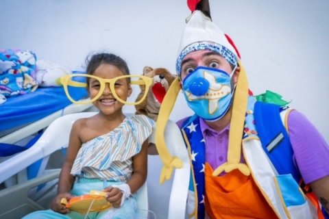 Ett barn bredvid en clown i sjukhusmiljö. Clownen håller upp ett par enorma solglasögon framför barnet som ler stort.