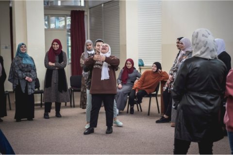 Ranin under en workshop. Hon står i en cirkel tillsammans med en annan kvinna som skrattar stort. Runt dem står andra kvinnor.