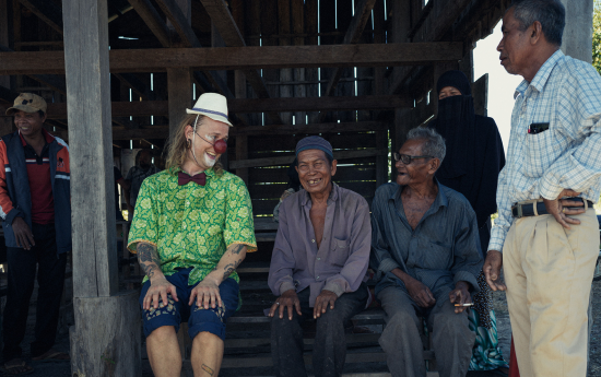 En clown i röd näsa, hatt, fluga och grönmönstrad skjorta sitter bredvid två äldre herrar. De tittar på varandra och skrattar.